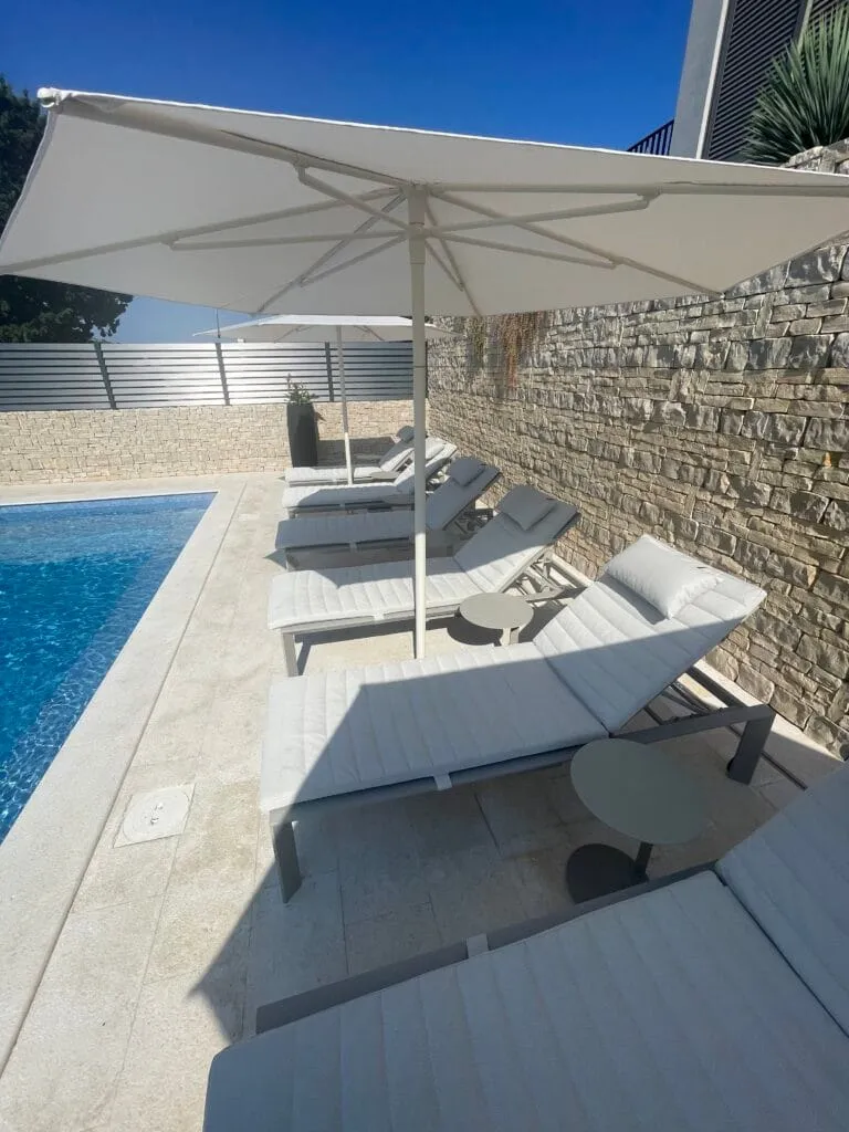 Bílé slunečníky Nata nad lehátky u bazénu lemovaného kamennou zdí v Chorvatsku